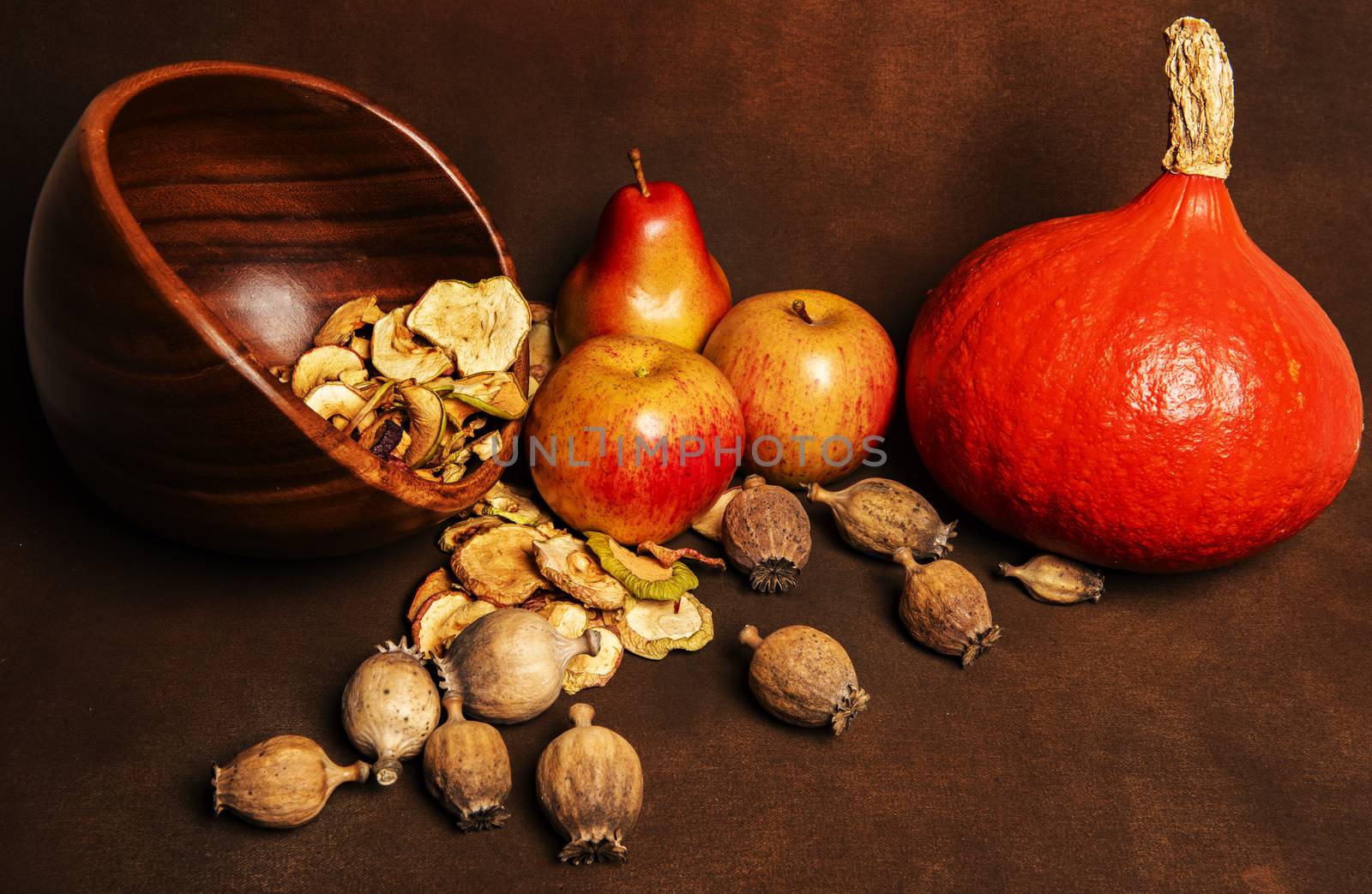 Still life of fresh red kuri squash, poppy heads and a pile of dried apples in a wooden bowl together with two whole apples and one pear. Still life concept of fall season harvest and homemade fruit processing ona brown background