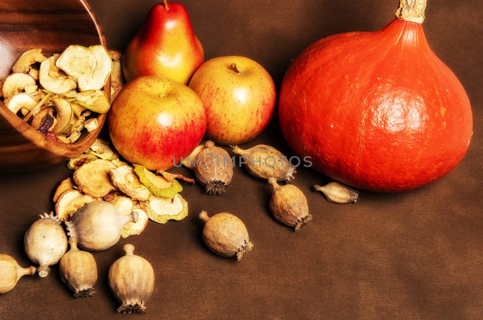 Still life of fresh red kuri squash, poppy heads and a pile of dried apples in a wooden bowl together with two whole apples and one pear. Still life concept of fall season harvest and homemade fruit processing ona brown background