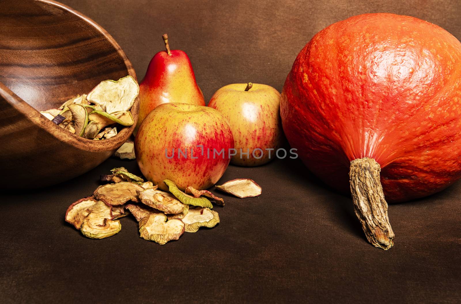 Still life of fresh red kuri squash, poppy heads and a pile of dried apples in a wooden bowl together with two whole apples and one pear. Still life concept of fall season harvest and homemade fruit processing ona brown background