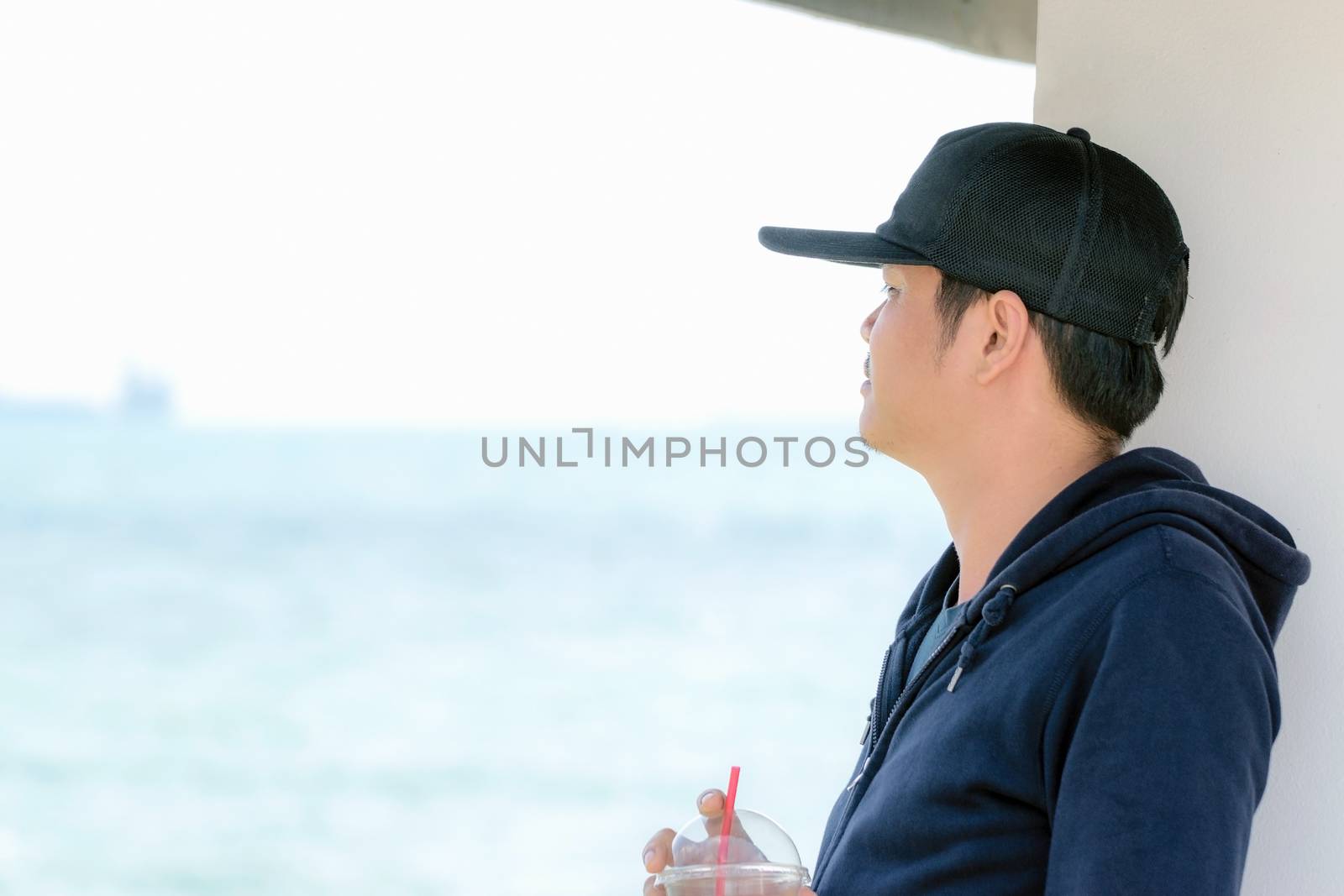 Asian man wearing a blue long sleeve shirt and wearing a hat holding a plastic glass of water by the ocean