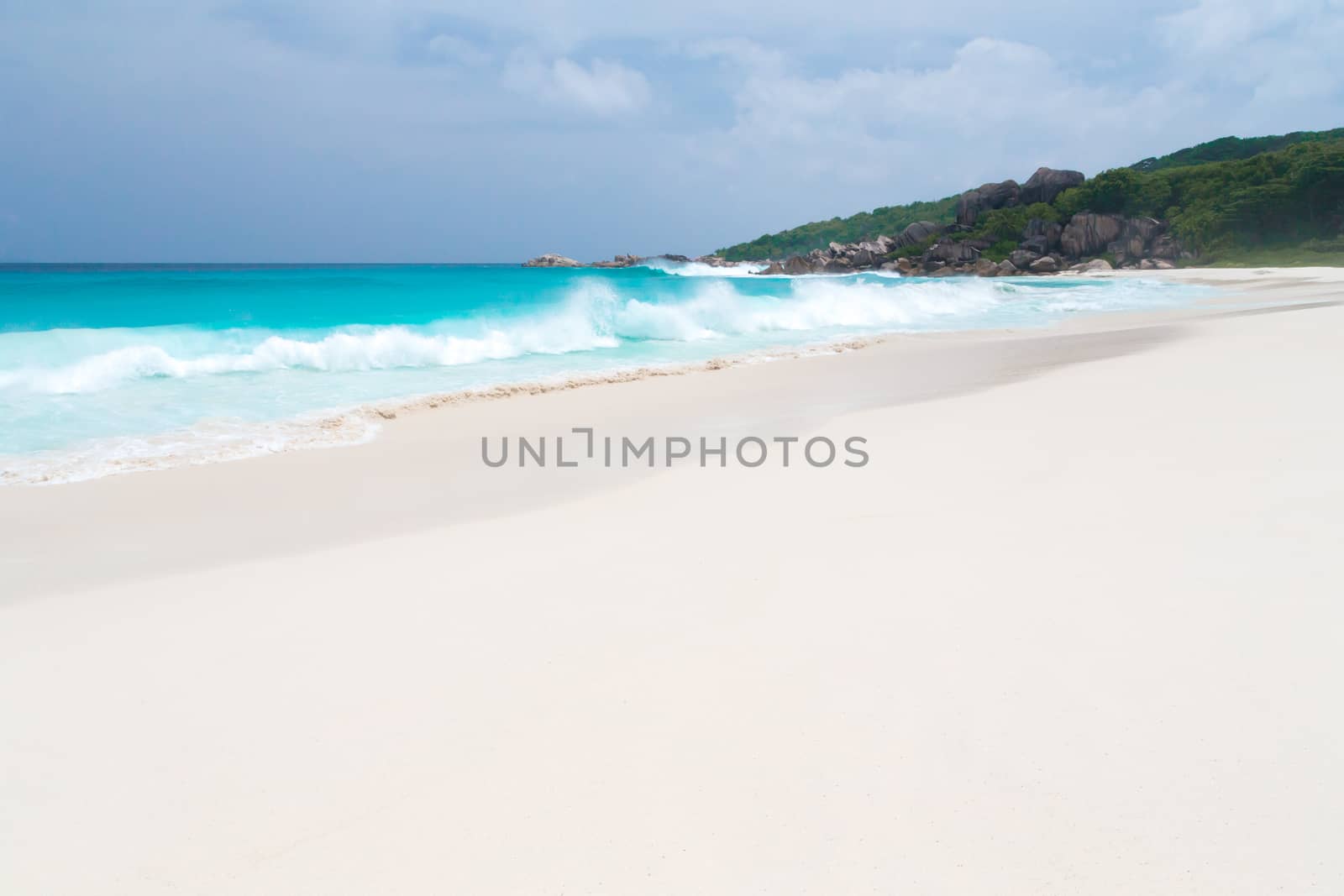 White sand beach with some waves and blue sky in the far a hill with green plants