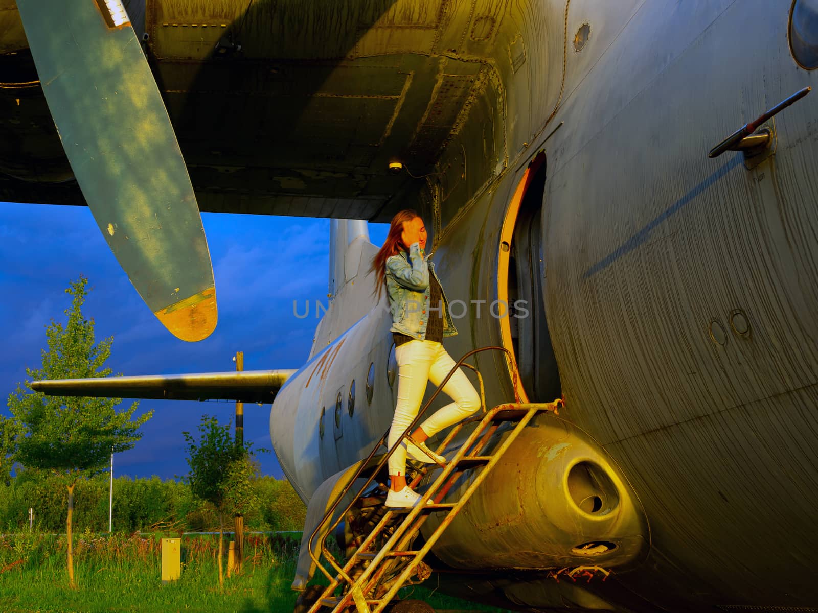 Yo ungwoman standing at plane ladder going to board, outdoors, airport. Old Soviet military airplane, sunset time. Close up of a Abandoned Historic AircraftAN-12. Close up of propeller engine.