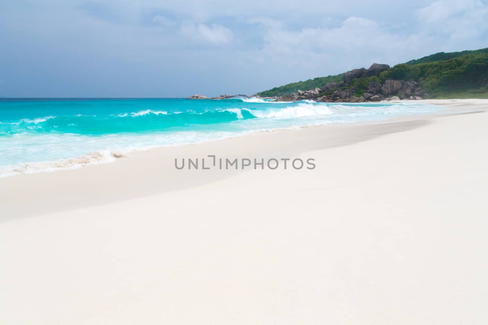 White sand beach with some waves and blue sky in the far a hill with green plants