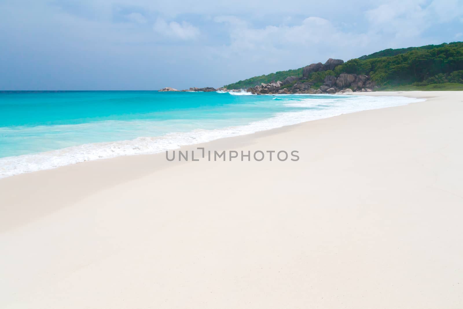 White sand beach with some waves and blue sky in the far a hill with green plants