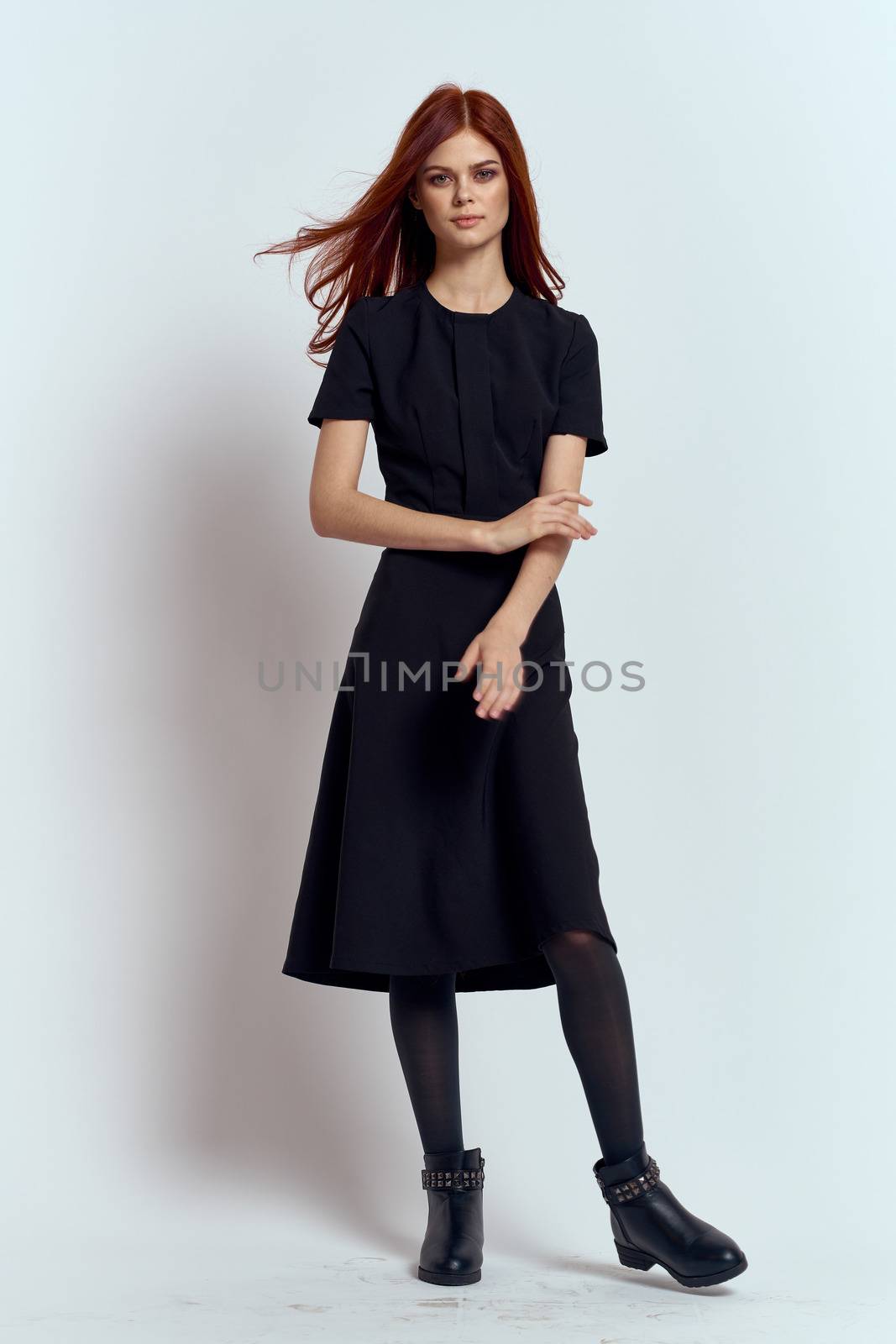 A woman in a black dress on a light background and pantyhose shoes red hair and pose in full growth. High quality photo