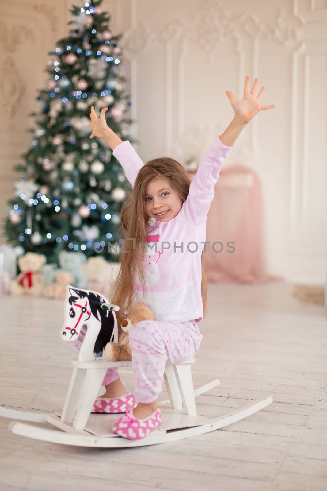 A beautiful little girl in pink pajamas rejoices in a wooden rocking horse, a gift from Santa for Christmas.