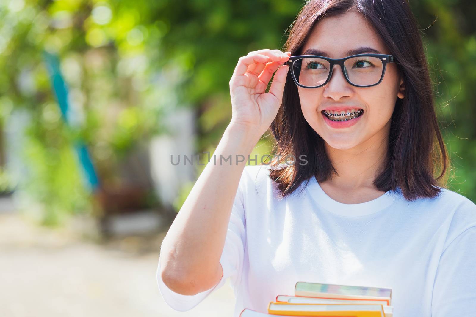 Portrait of Asian teen beautiful young woman wear eyeglasses smile have dental braces on teeth laughing at outdoor she hold education books on hand, Medicine and dentistry concept