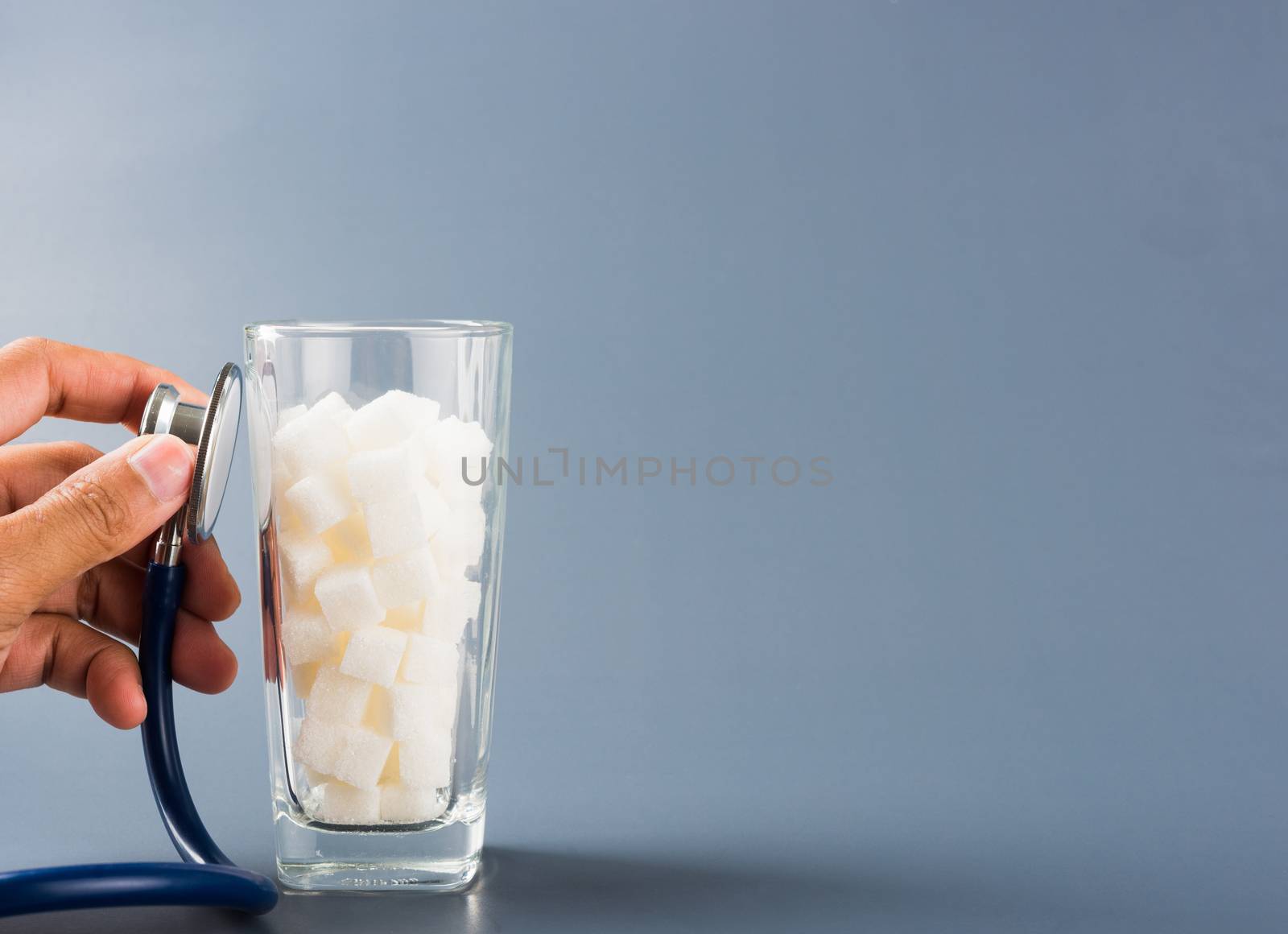 Hand of doctor hold stethoscope check on glass full of white sugar cube sweet food ingredient, isolated on gray background, health high blood risk of diabetes and calorie intake unhealthy drink