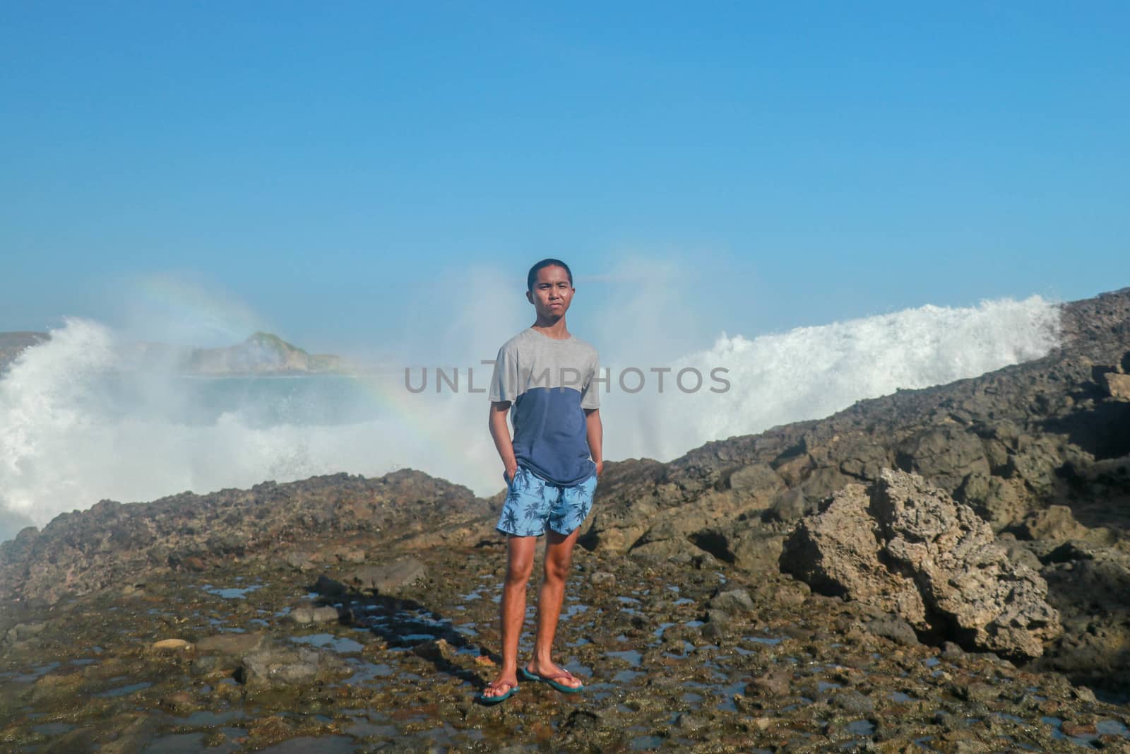 Waves hitting round rocks and splashing. A young man stands on a rocky shore and the waves crash against a cliff.