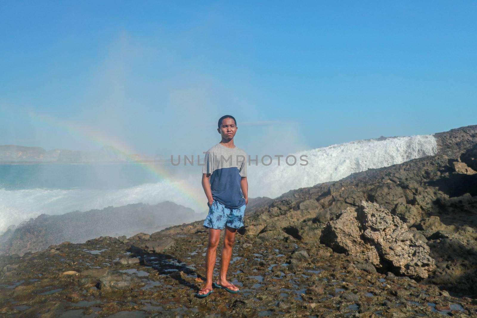 A man stands on a rocky shore and the waves crash against a cliff. Rainbow phenomenon in water fog. Waves hitting round rocks and splashing by Sanatana2008