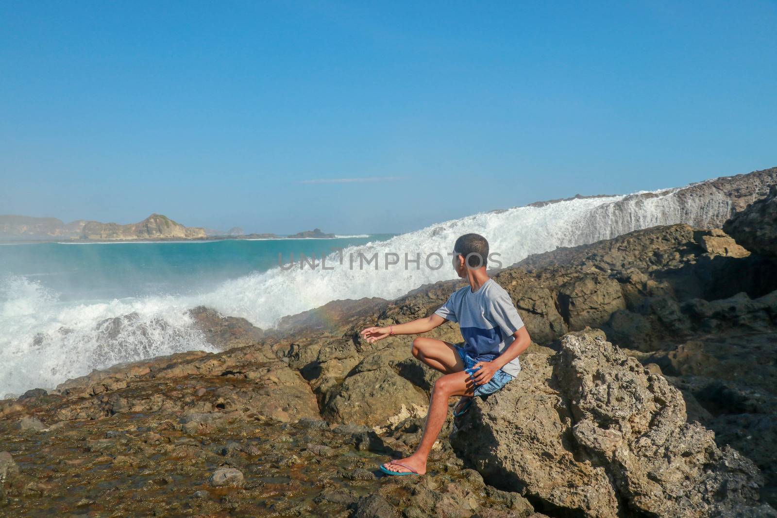 young man in a T-shirt and blue shorts sitting on a rocky shore. This is a view of man sitting on the rock by the sea.