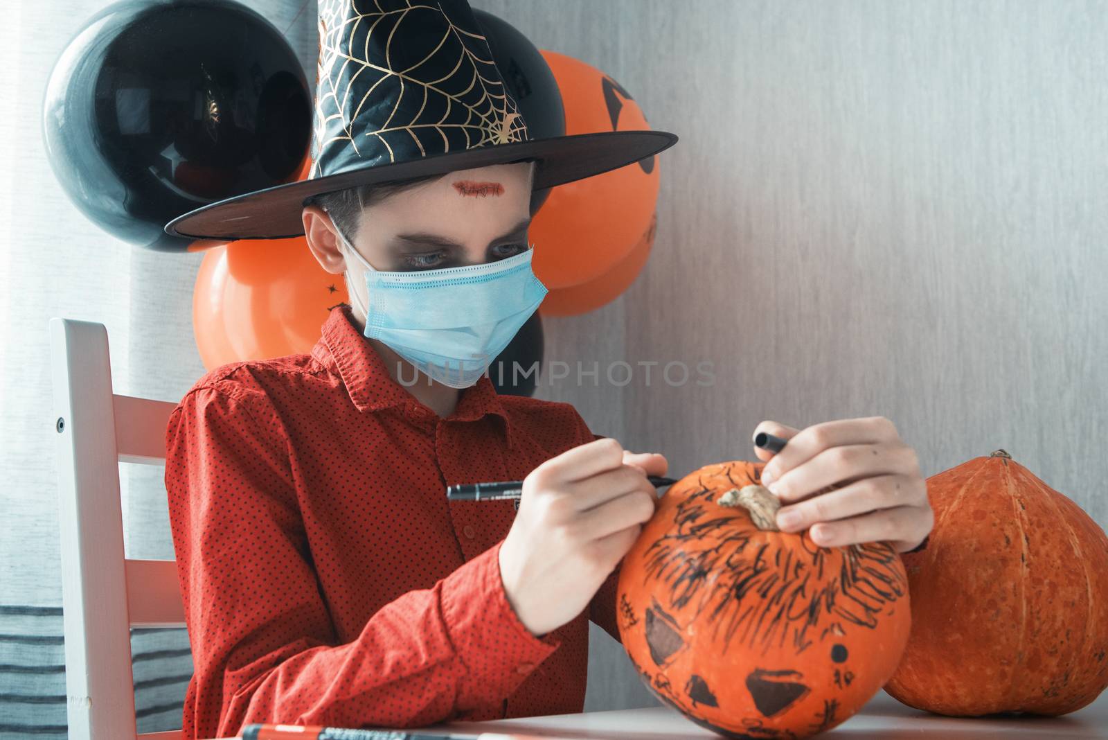 Teen boy in costume and face masks to protect against COVID-19 drawing a pumpkin for the Halloween celebration. Halloween carnival with new reality with pandemic concept.