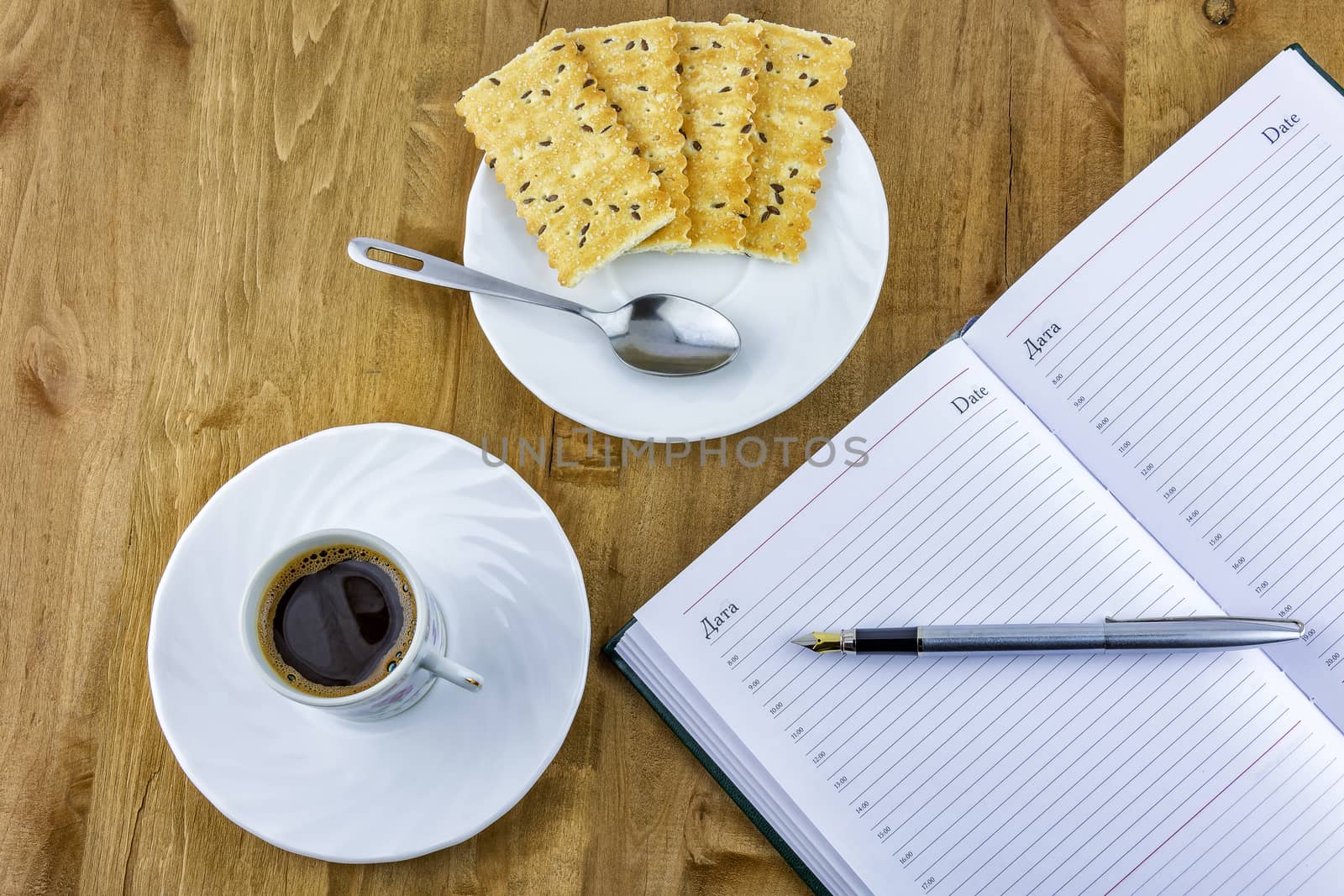 On a wooden surface is a cup of coffee on a saucer and biscuits and is open daily with a pen