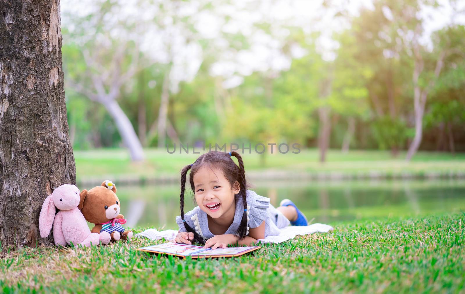 cute little girl  reading a book while lying with a doll in the park