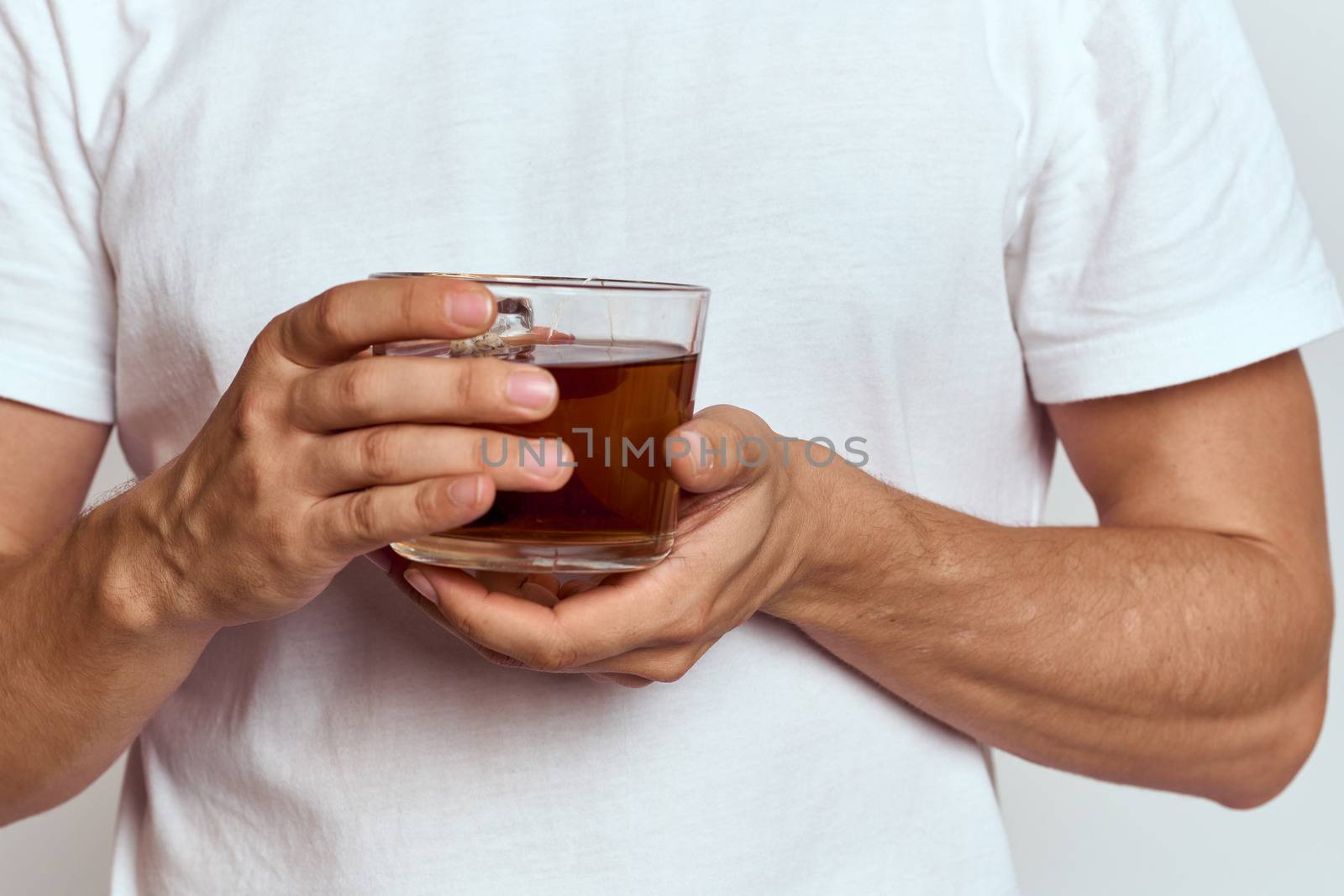 A man with a hot drink of tea in his hands in a white T-shirt on a light background cropped view by SHOTPRIME