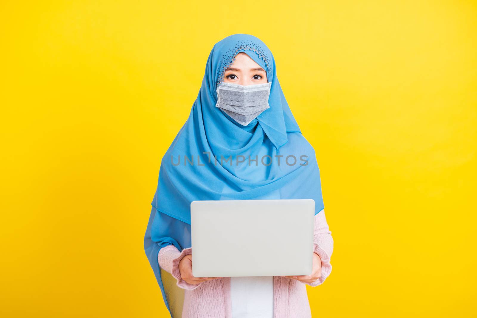 Asian Muslim Arab, Portrait of happy beautiful young woman religious wear veil hijab and face mask protective to prevent coronavirus she holding the laptop typing, isolated on yellow background