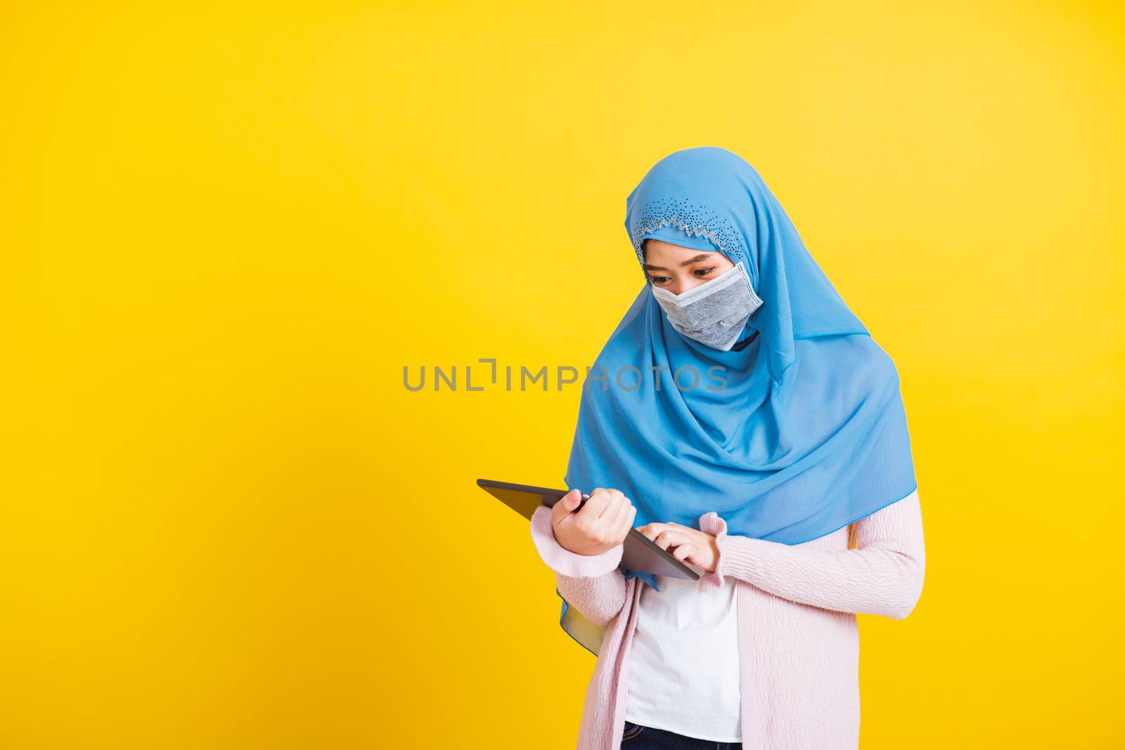 Asian Muslim Arab, Portrait of happy beautiful young woman Islam religious wear veil hijab and face mask protect she quarantines disease coronavirus touch on tablet screen isolated yellow background