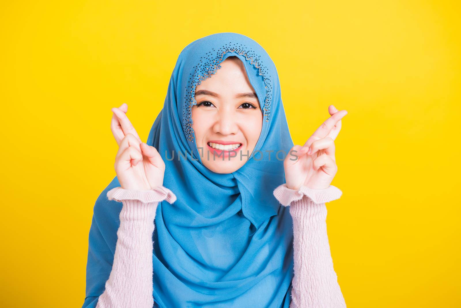 Asian Muslim Arab, Portrait of happy Asian beautiful young woman Islamic religious wear veil hijab she smiling and holding fingers crossed for good luck, studio shot isolated on yellow background