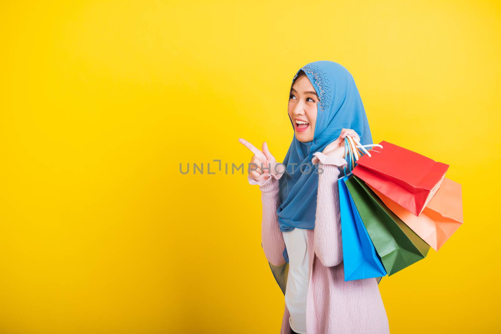 Asian Muslim Arab, Portrait of happy beautiful young woman Islam religious wear veil hijab funny smile she holding colorful shopping bags, studio shot isolated on yellow background