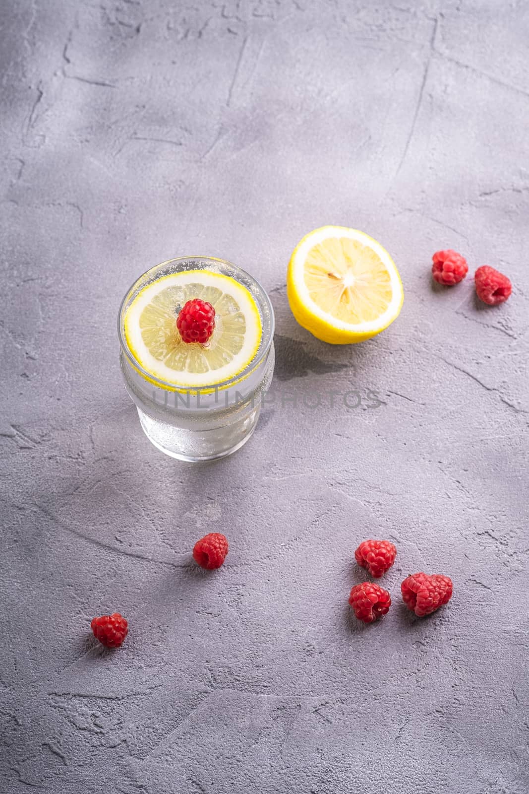 Fresh cold sparkling water drink with lemon, raspberry fruits in glass on stone concrete background, summer diet beverage, angle view selective focus