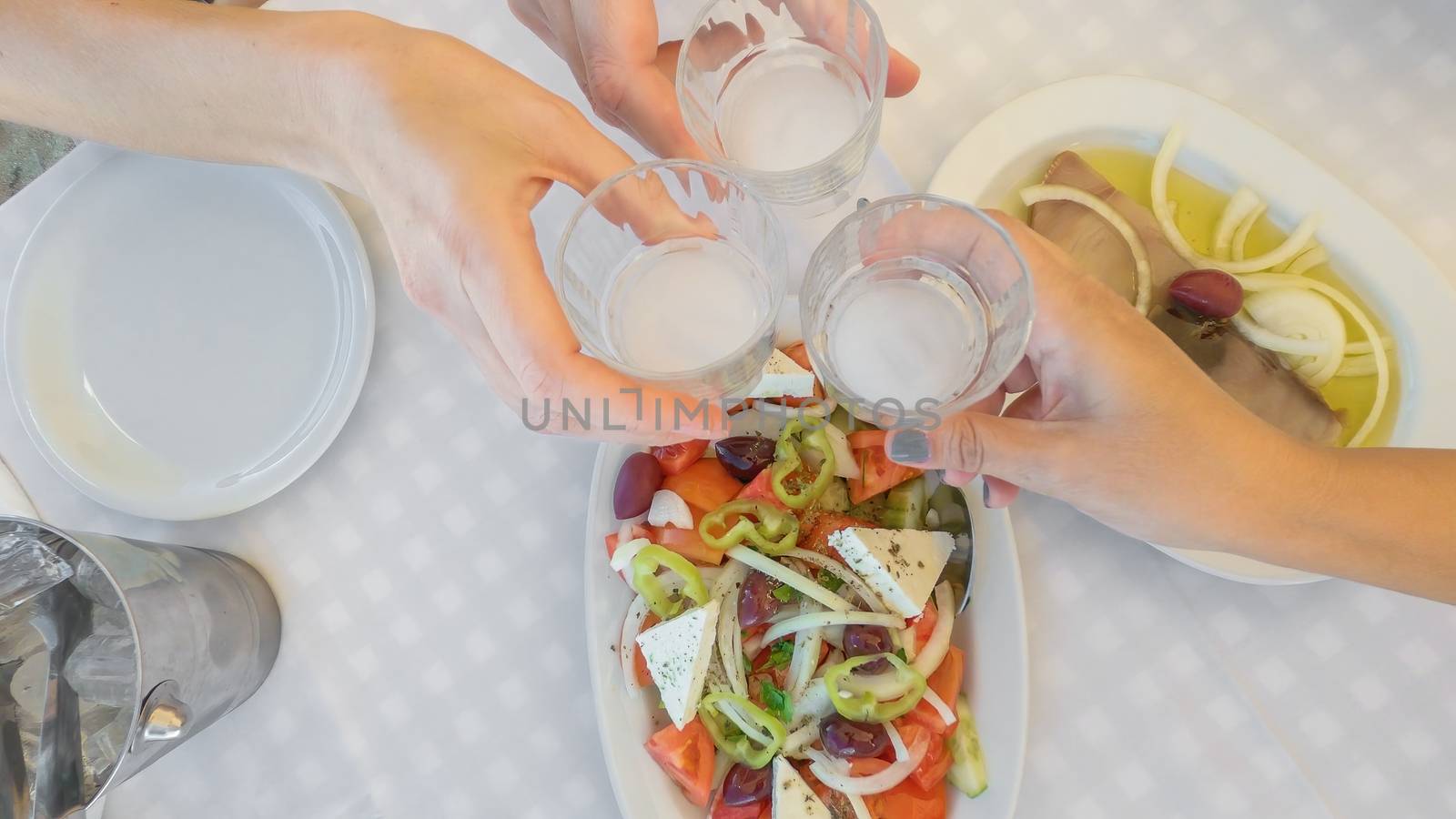 Social bonding with people clinking with glasses of alcoholic ouzo drink above a tavern table with Greek salad and fish meze starter food.
