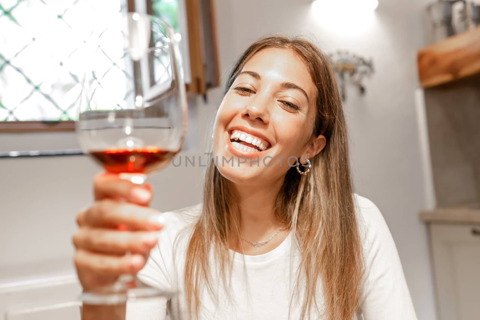 Cute young caucasian blonde woman toasting with red wine glass looking at the camera at home - New normal remote conference communication by online technology for the Coronavirus pandemic by robbyfontanesi