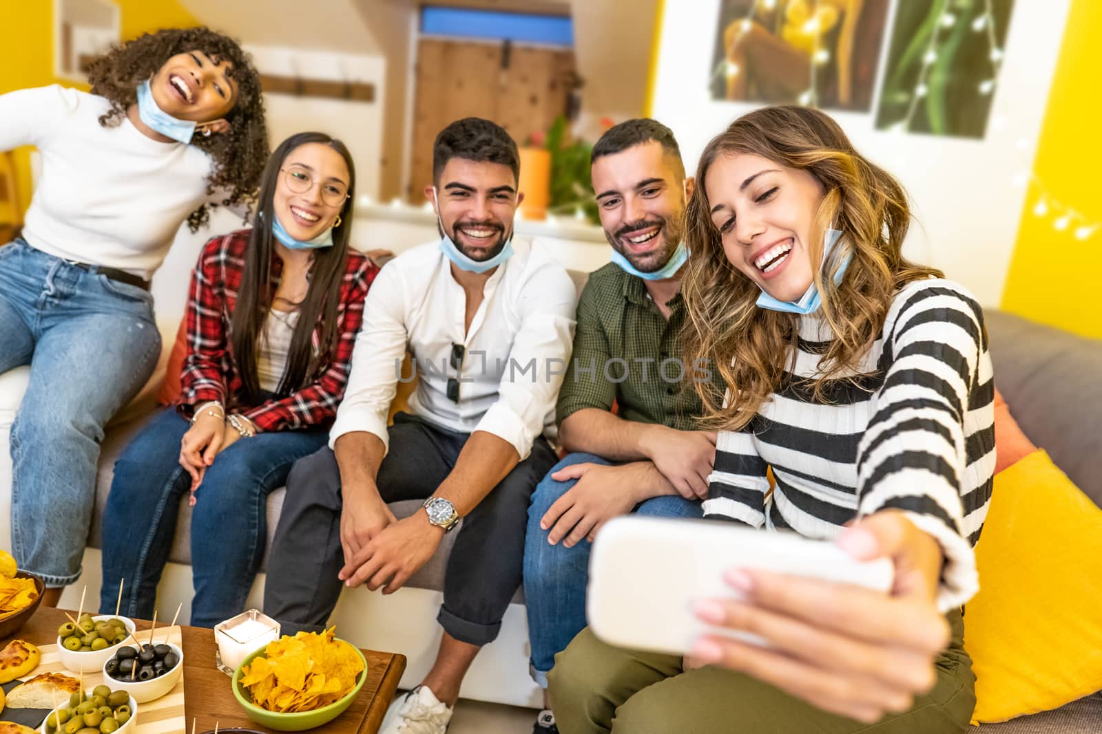 Group of mixed race friends making selfie celebrating at home with medical protection mask - Happy people indoor aperitif party at the Coronavirus new normality time by robbyfontanesi