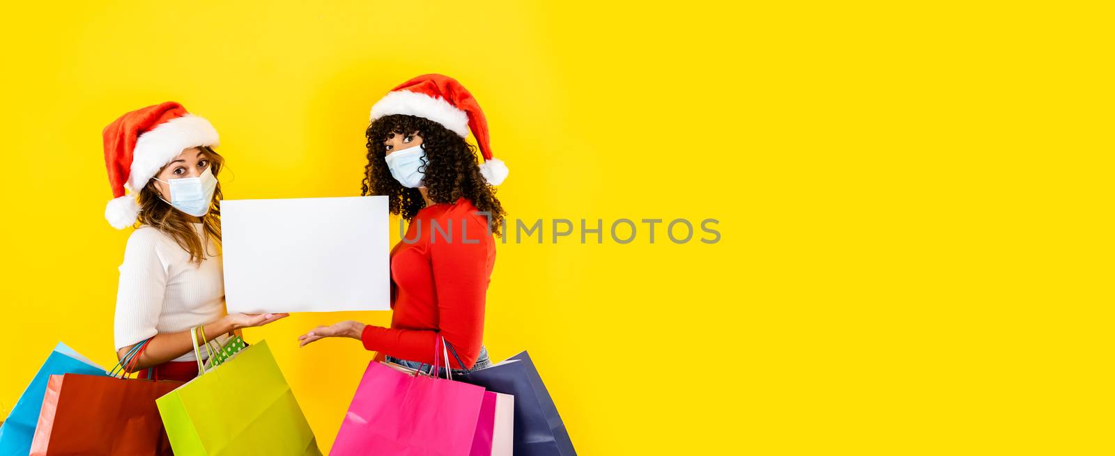Two young cute women wearing Santa Claus hat looking to the camera with wide eyes and medical mask, holding sign for copy space with many shopping bags in arms - Christmas banner for Coronavirus by robbyfontanesi