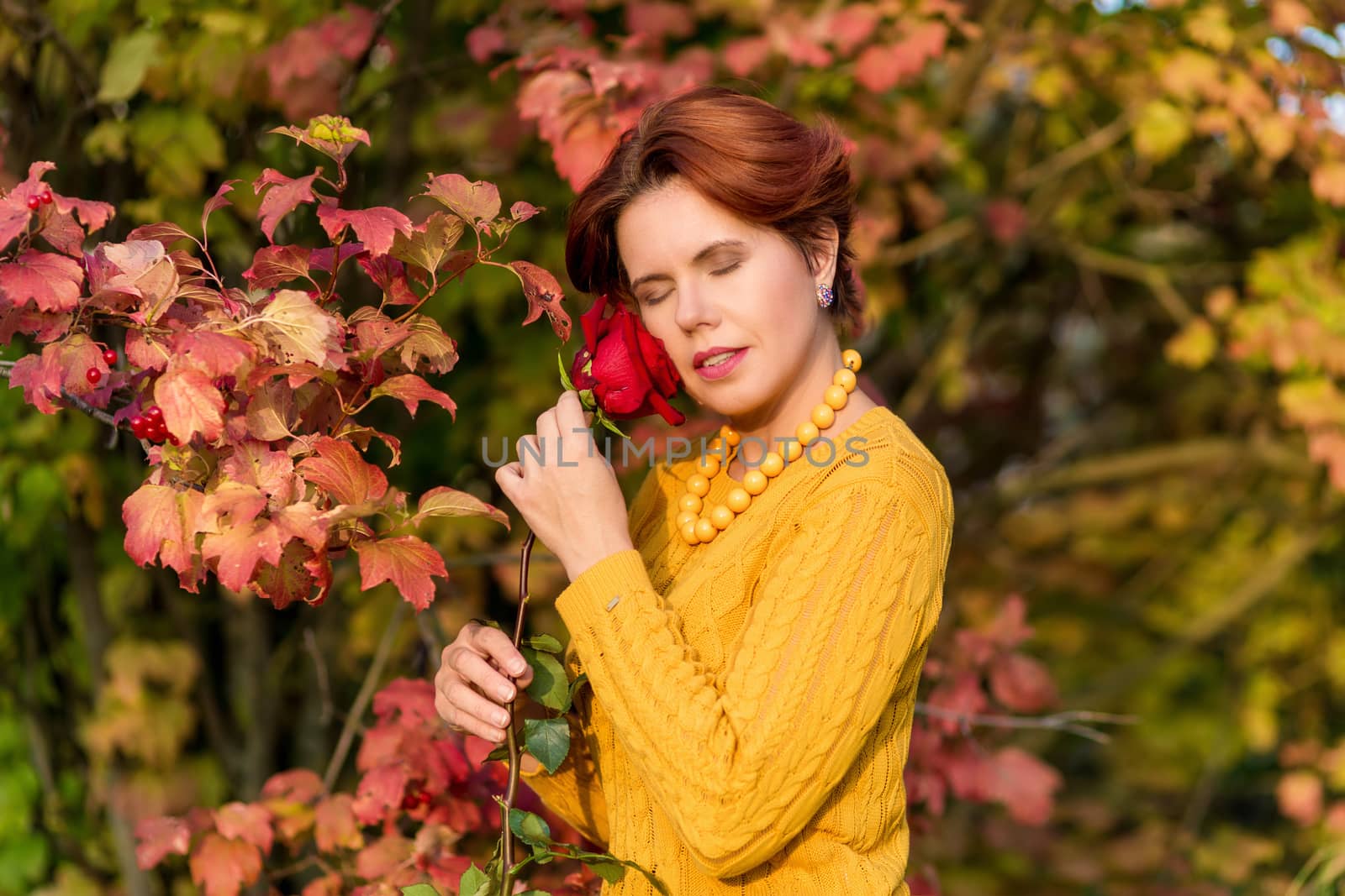 Portrait of beautiful 30 year old woman holding rose and standing near Red foliage on a viburnum Bush with closed eyes in autumn park
