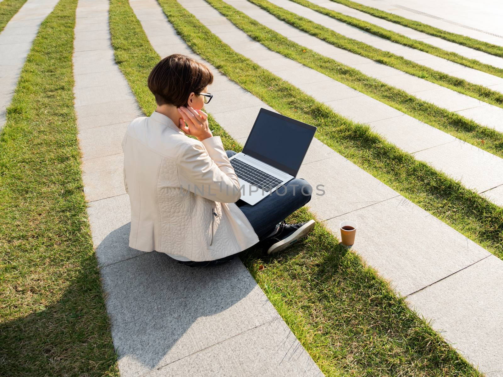 Freelance business woman sits in park with laptop and take away cardboard cup of coffee. Student studies online. Casual clothes, urban lifestyle of millennials.