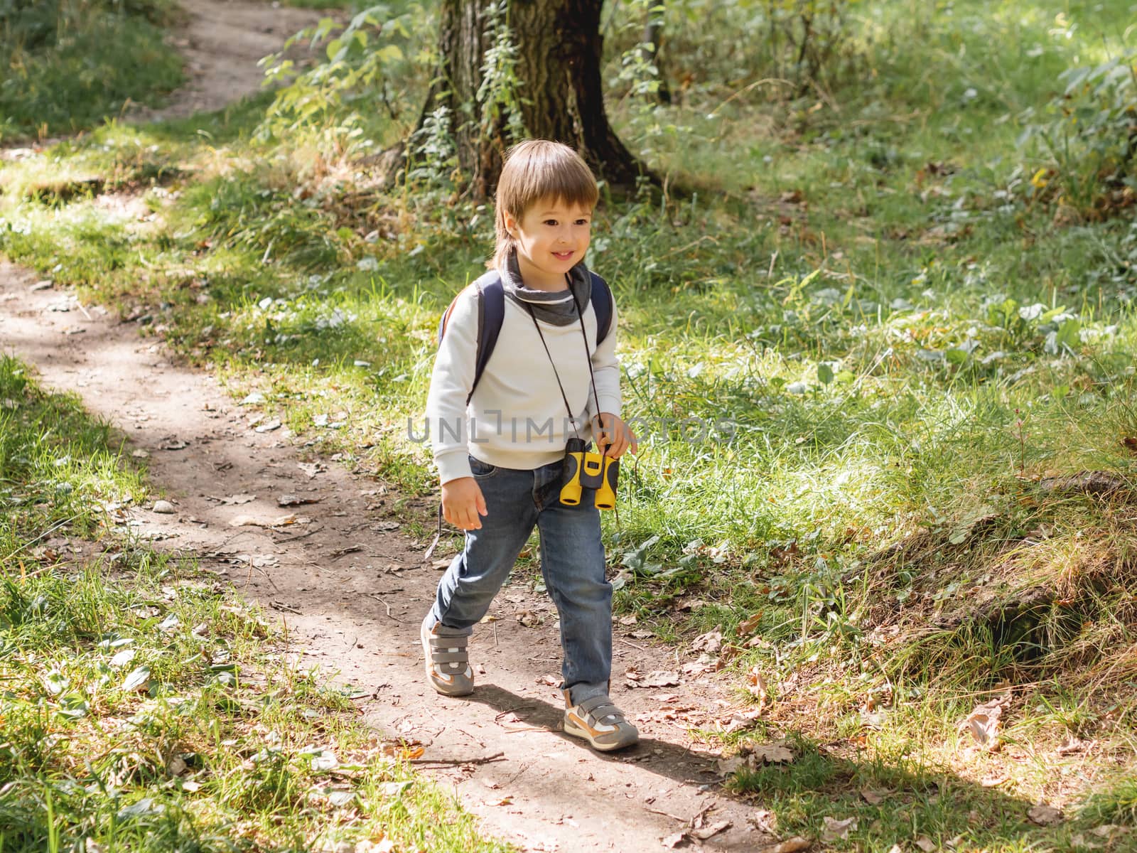 Curious boy is hiking in forest lit by sunlight. Outdoor leisure activity for kids. Child with binoculars and backpack.