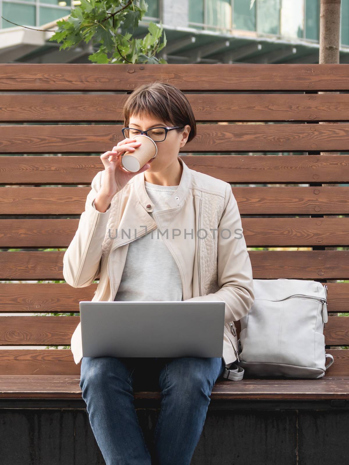 Freelance business woman sits in park with laptop and take away cardboard cup of coffee. Casual clothes, urban lifestyle of millennials. Working remotely.
