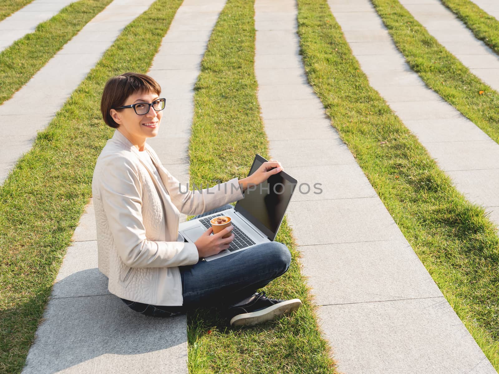 Business woman sits in park with laptop and take away cardboard cup of coffee. Casual clothes, urban lifestyle of millennials.