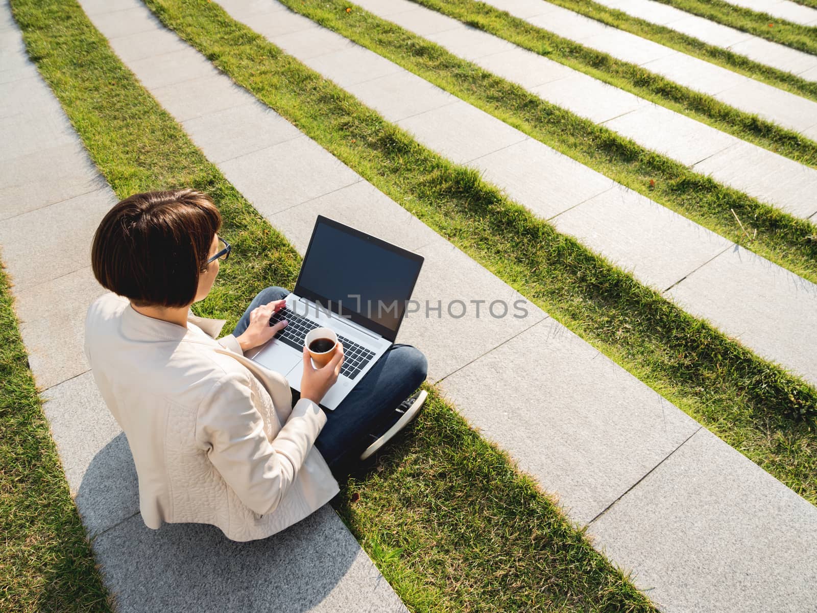 Business woman sits in park with laptop and take away cardboard by aksenovko