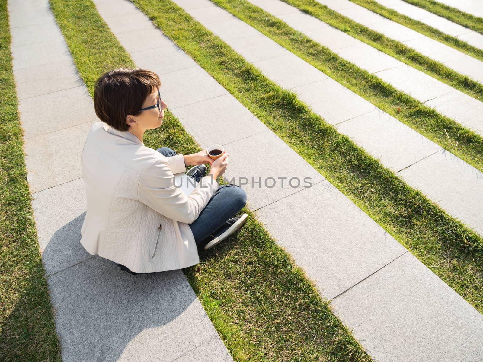 Woman with short haircut and eyeglasses sits in park with take away cup of coffee. Woman enjoys summer sun. Casual clothes, urban lifestyle of millennials.