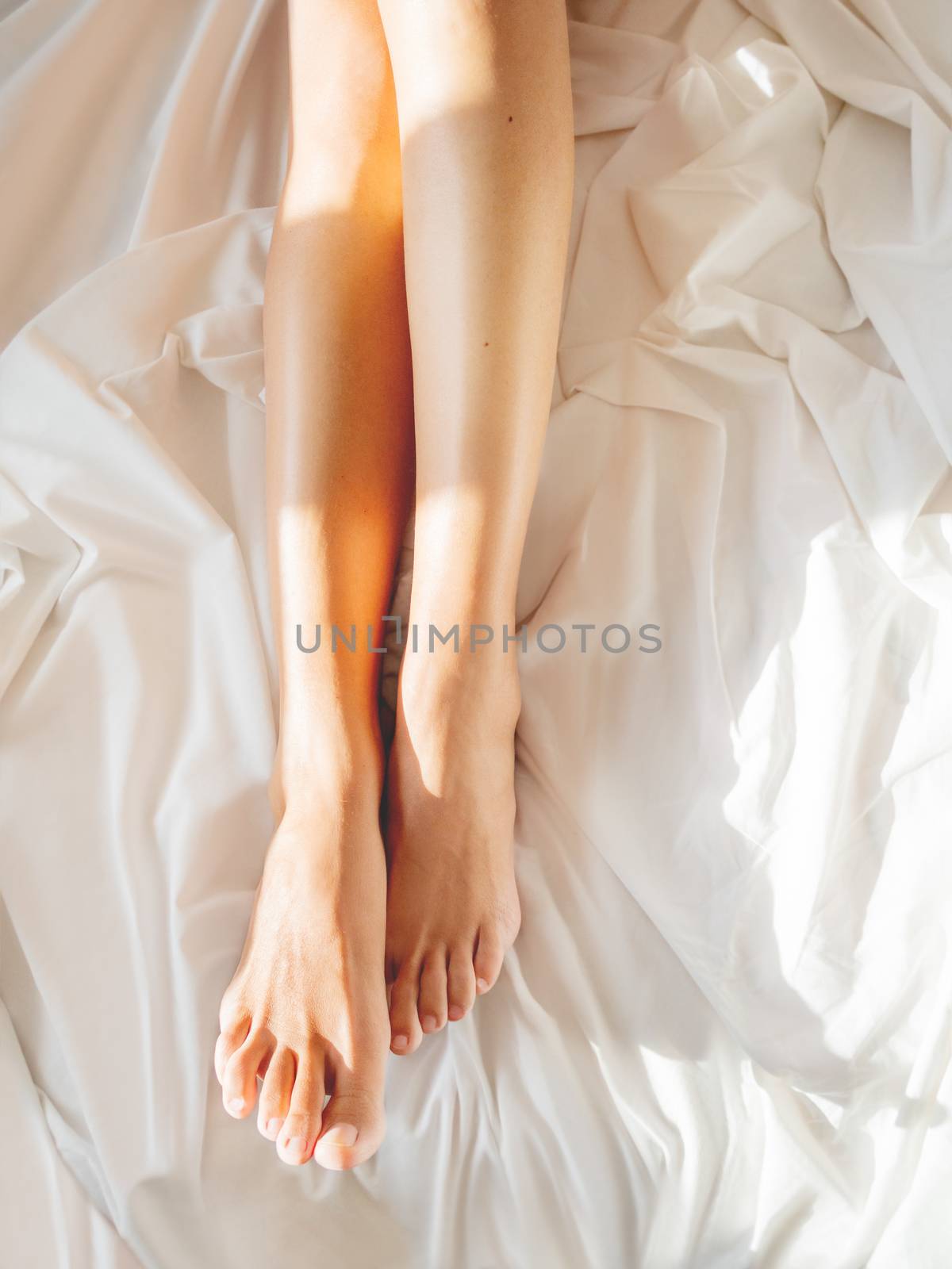 Morning bedtime. Woman's feet on unmade bed. Sunbeams on white c by aksenovko