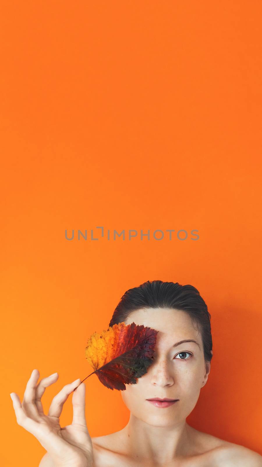 Portrait of Asian woman closing her eye with autumn leaf. Model on orange monochrome background. Natural beauty on bright and colorful backdrop. Banner with copy space.