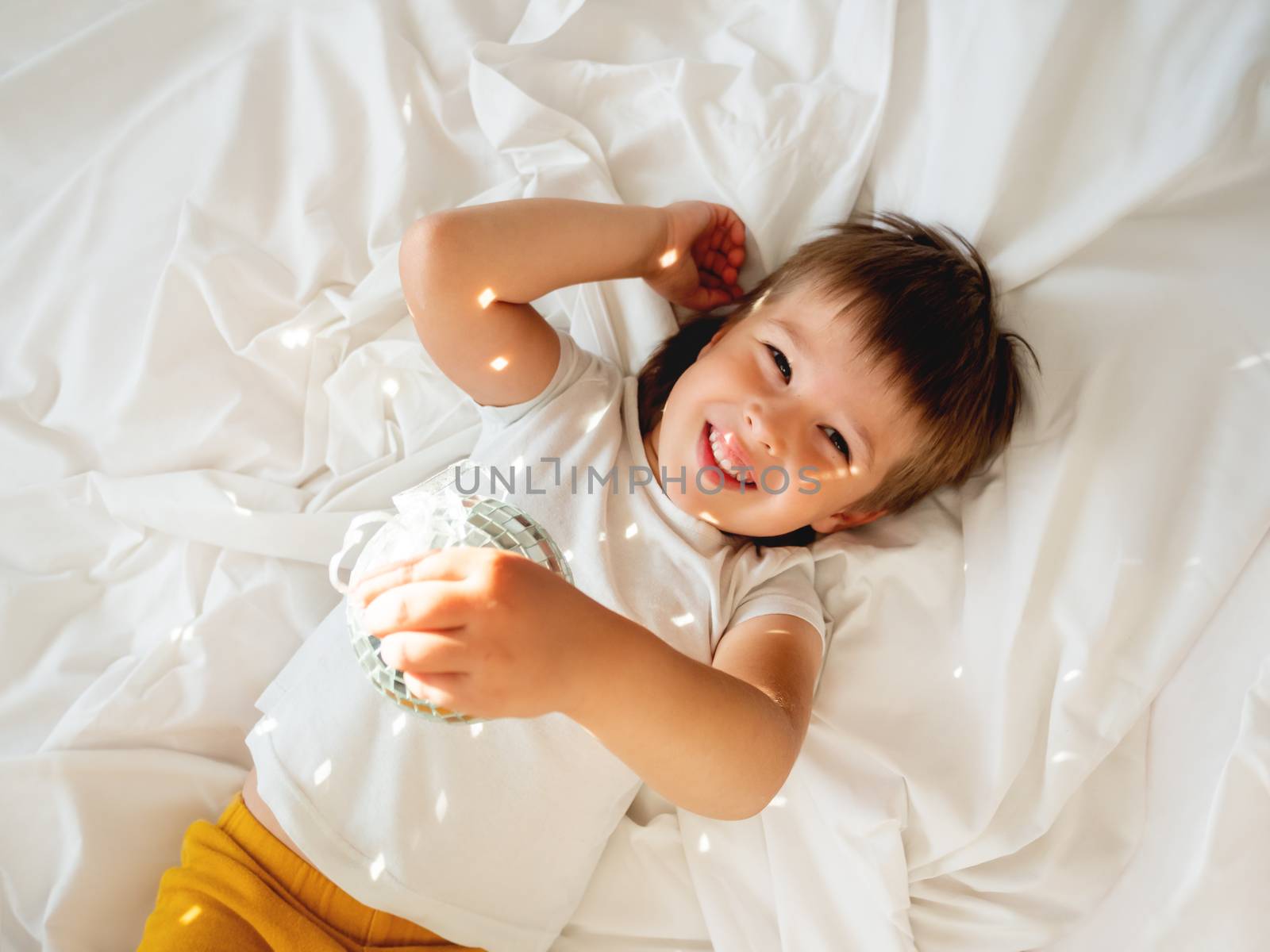 Little boy plays with sparkling mirror disco ball in bed and laughing happily. Joyful toddler. Playful child and sunny morning in cozy home.