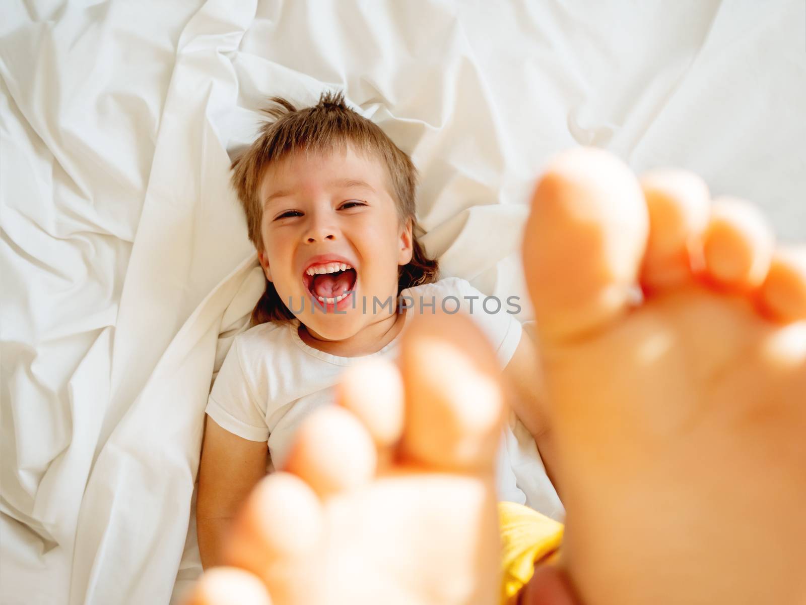 Little boy is lying upside down on the bed and laughing happily. Joyful toddler. Playful child shows his feet in the air. Sunny morning in cozy home.