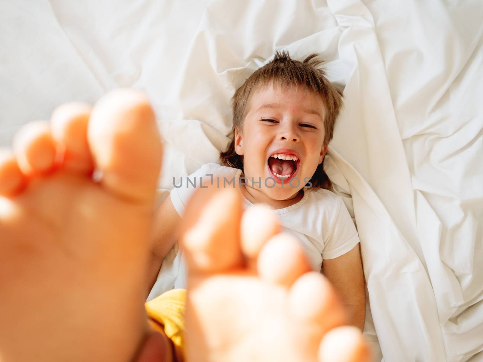 Little boy is lying upside down on the bed and laughing happily. Joyful toddler. Playful child shows his feet in the air. Sunny morning in cozy home.