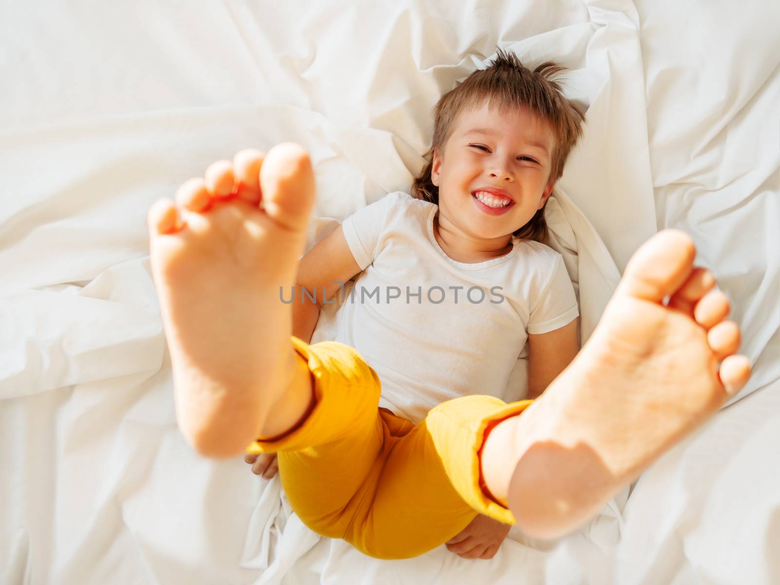 Little boy is lying upside down on the bed and laughing happily. Joyful toddler. Playful child shows his feet in the air. Sunny morning in cozy home.