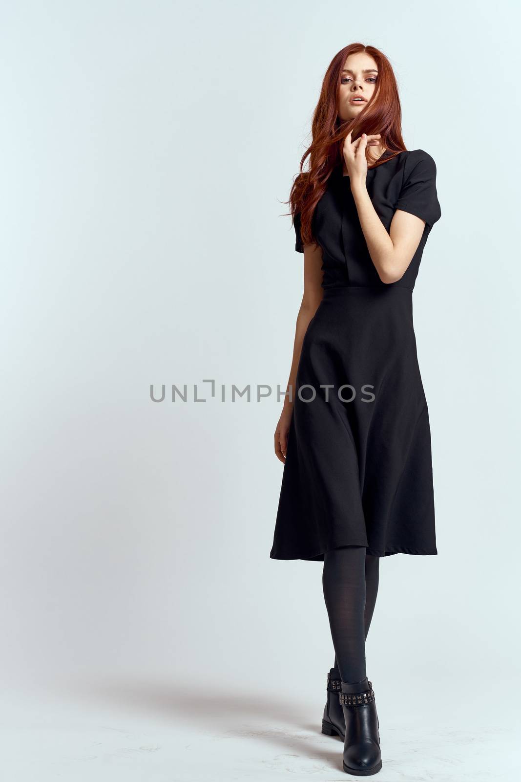 A woman in a black dress on a light background and pantyhose shoes red hair and pose in full growth by SHOTPRIME