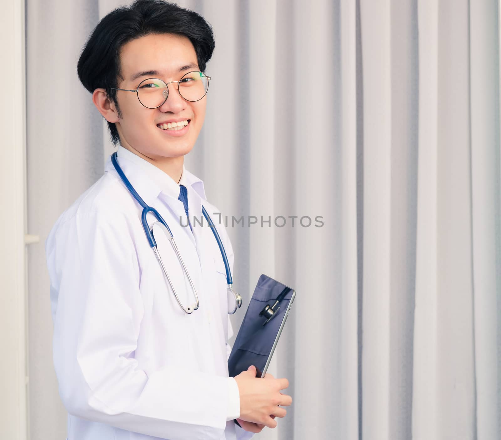 Portrait closeup of Happy Asian young doctor handsome man smiling in uniform and stethoscope neck strap holding smart digital tablet on hand and looking to the camera, healthcare medicine concept