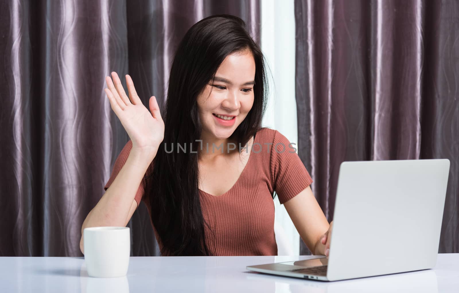 Work from home, Smiling Happy Asian business young beautiful woman sit on desk workspace video call conferencing by webcam laptop computer technology and raise your hand and say hello at home office