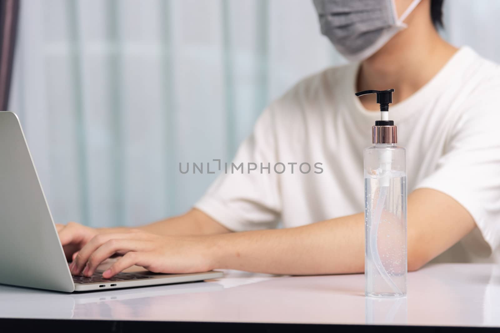 Asian business young handsome man glasses, wearing protective mask with disinfectant gel beside he work from home office remote online with laptop computer quarantines disease coronavirus or COVID-19