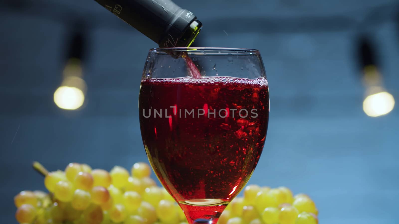 Close up red wine pouring into a glass. Bunches of grapes on the table. Blurry lamps on blue background