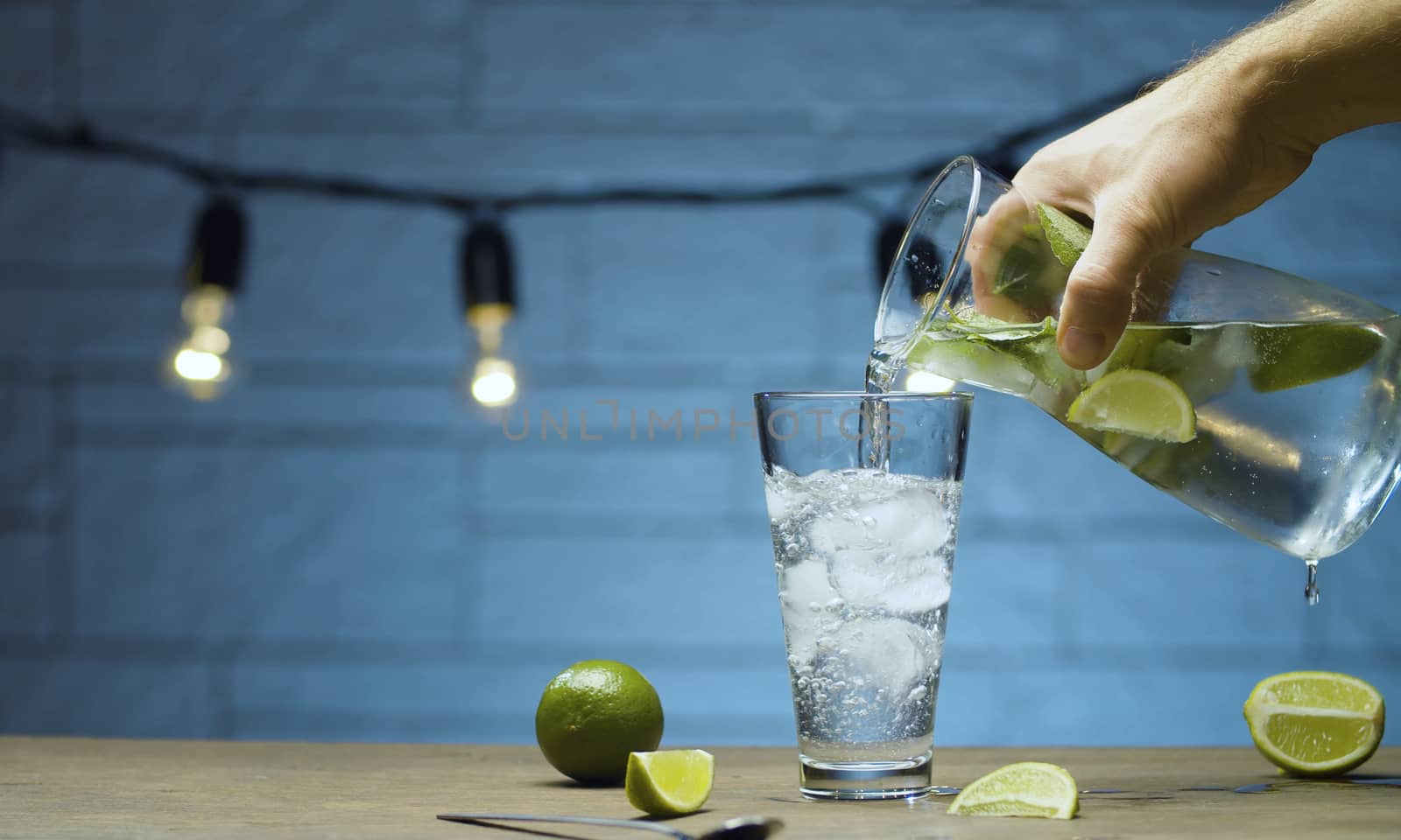 Close up male hand pouring lemonade with lime and mint into a glass. Blurry lamps on blue background