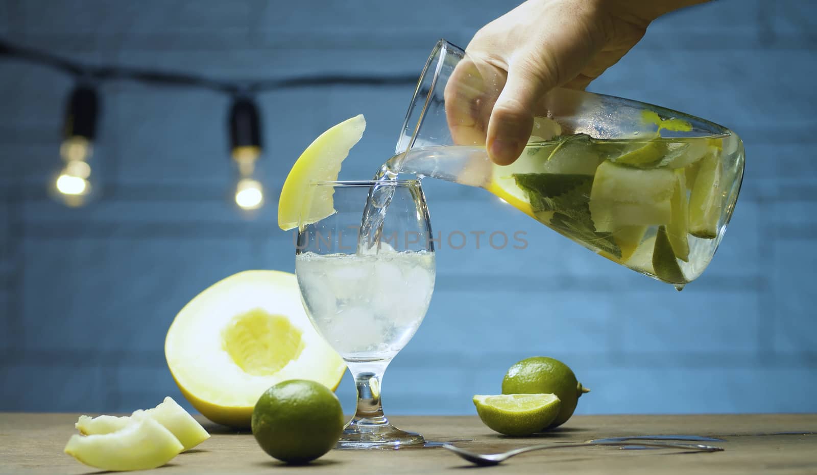 Close up male hand pouring melon lemonade into a glass from a jug with pieces of melon, lime wedges, mint leaves and ice. Blurry lamps on blue background