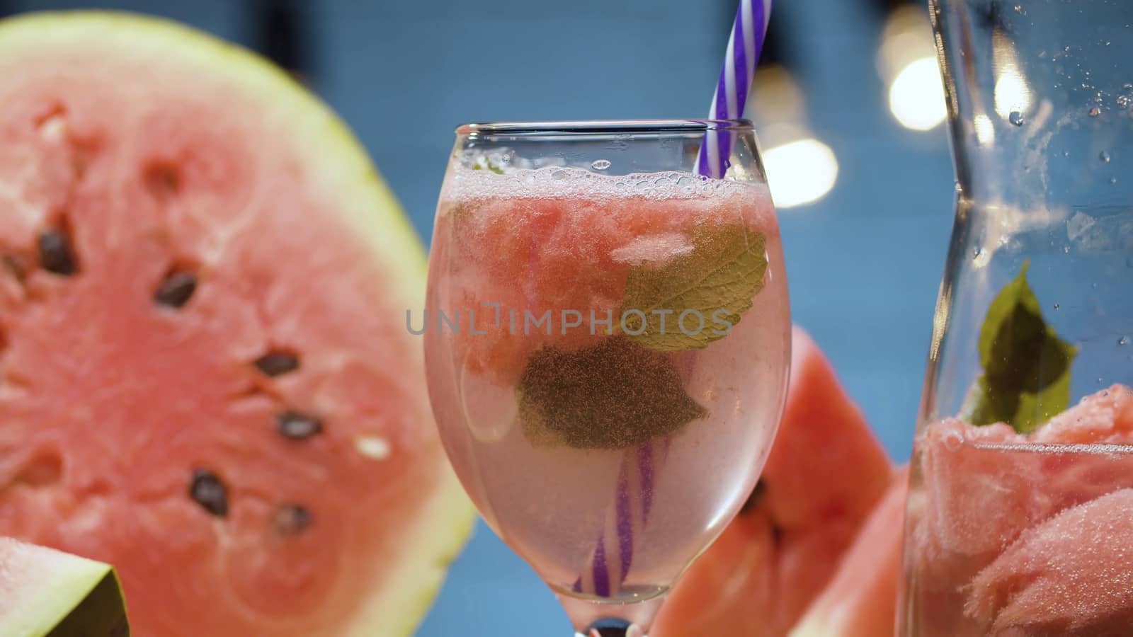 Close up watermelon lemonade in a glass. Soda water, pieces of watermelon, mint leaves and ice in glass jug. Blurry lamps on blue background