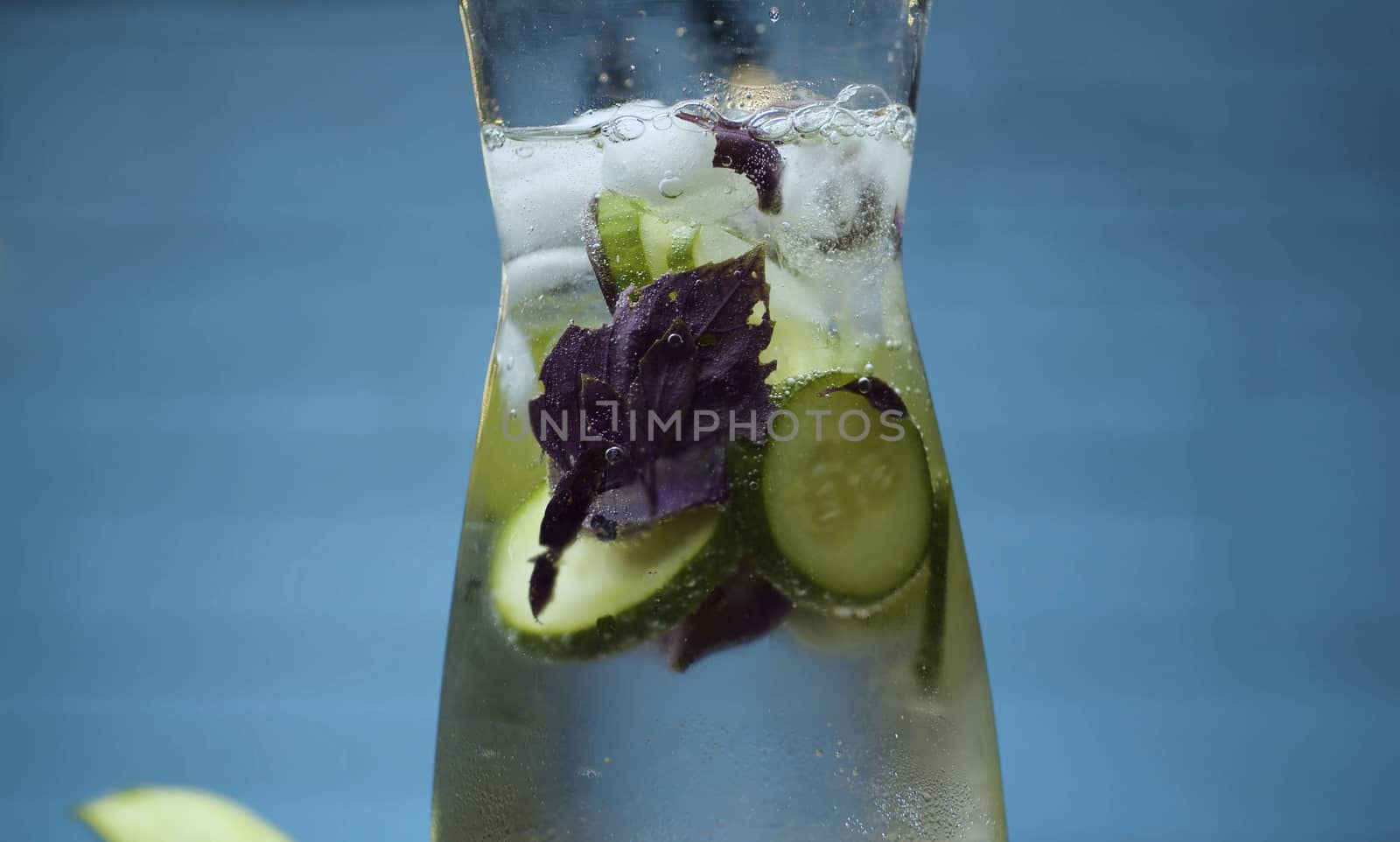 Close up vegetable fizzy drink. Sparkling water in a glass jug with sliced cucumber and purple basil leaves. Blurry bulbs on blue background